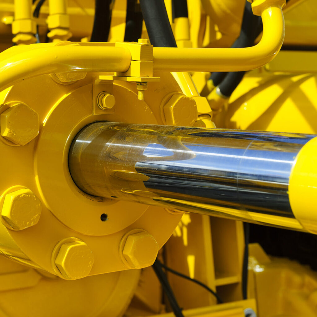 Close-up of a shiny metal hydraulic piston rod attached to yellow industrial machinery, with bolts and hydraulic hoses visible in the background.