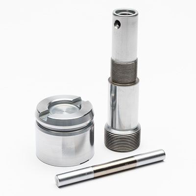 Three precision-machined metal parts—one cylindrical rod, one threaded shaft with holes, and one round component with a recessed top—are arranged on a white background.