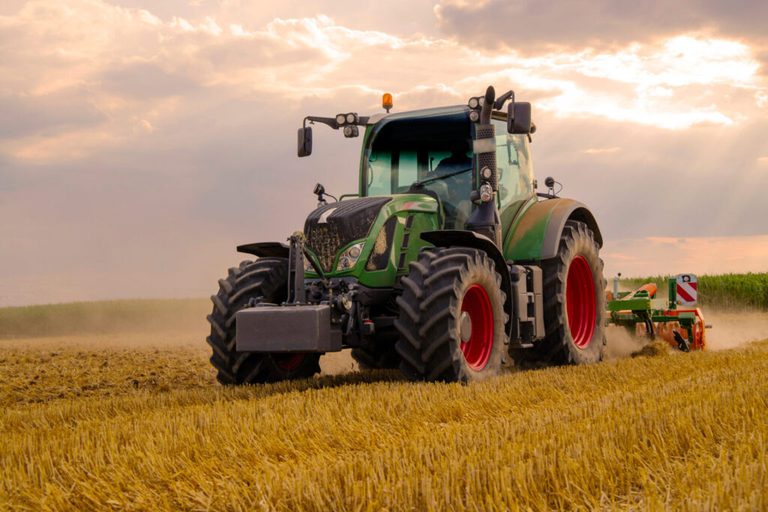 A green tractor with large tires plows a golden field under a partly cloudy sky, with dust rising behind it. Cornfields are visible in the background.