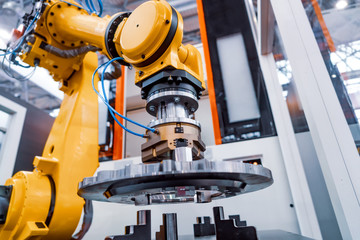 A yellow industrial robot arm with a mechanical gripper is placing a metal component on a machine in a modern factory setting.