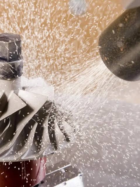 Close-up of a metal component being cleaned or processed with a high-pressure spray, causing water or particles to scatter in various directions.