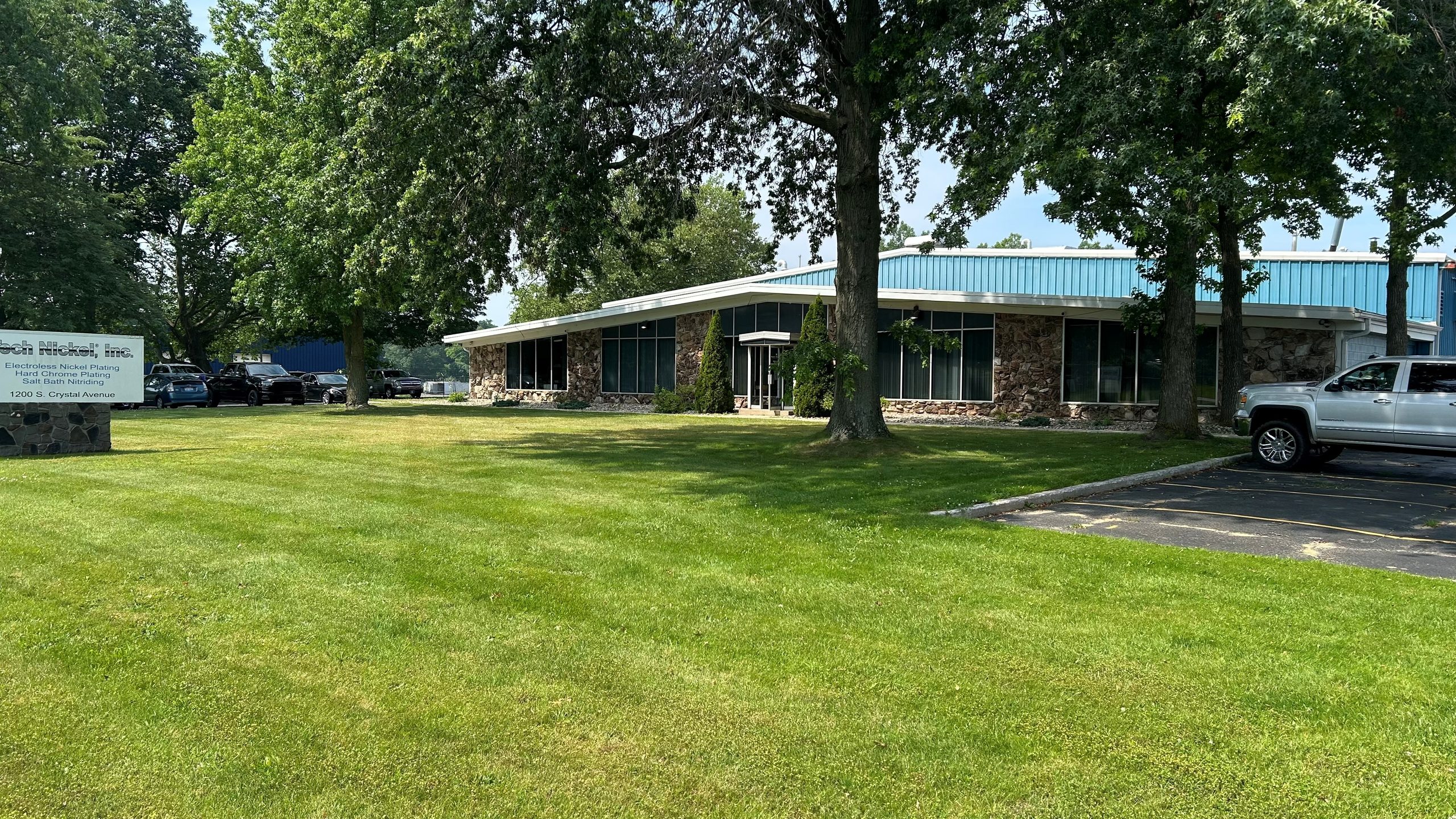 A single-story office building with large windows, surrounded by trees and grass. Several cars are parked nearby, and a sign is visible on the left side of the lawn.