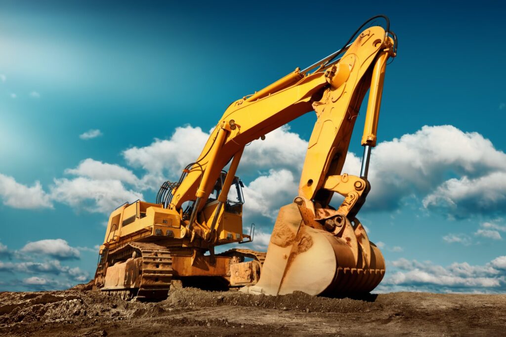 A large yellow excavator sits on a dirt construction site under a blue sky with scattered clouds, its bucket resting on the ground.