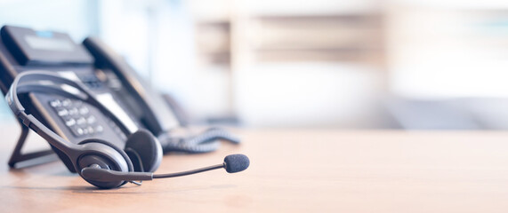 A headset with a microphone rests on a wooden desk next to an office phone, with a blurred background suggesting a workplace or call center environment.