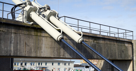 Two large hydraulic pistons are attached to a concrete bridge, positioned at an angle. Buildings and a blue sky with clouds are visible in the background.