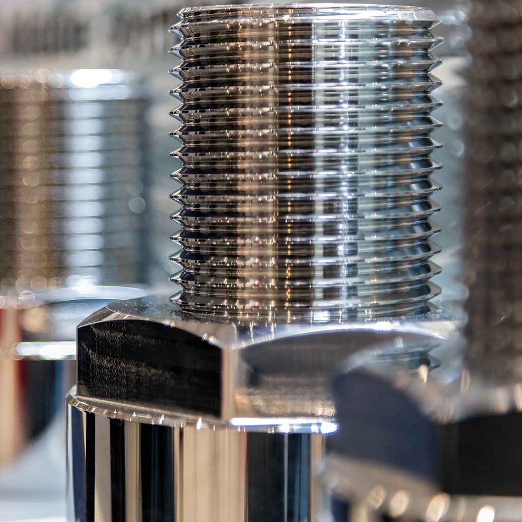 Close-up view of shiny metal bolts, with visible threading and hexagonal heads, arranged vertically in a row. The focus is on the central bolt, while others appear blurred in the background.