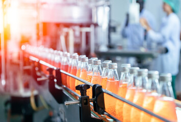 Clear glass bottles filled with orange liquid move along a conveyor belt in a factory. Workers in blue uniforms and caps are blurred in the background.