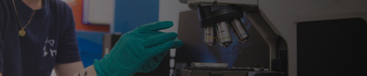 A person wearing a teal glove adjusts the lens of a microscope in a laboratory setting.