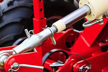 Close-up of hydraulic machinery parts, showing a metal hydraulic cylinder attached to red painted equipment. A large black tire is visible in the blurred background.