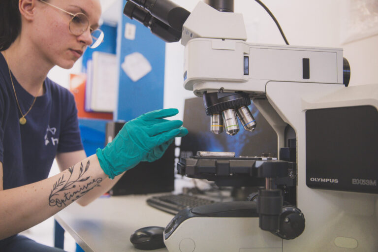 A person wearing glasses and teal gloves adjusts a sample under a microscope in a laboratory. A tattoo reading optimism is visible on their forearm.