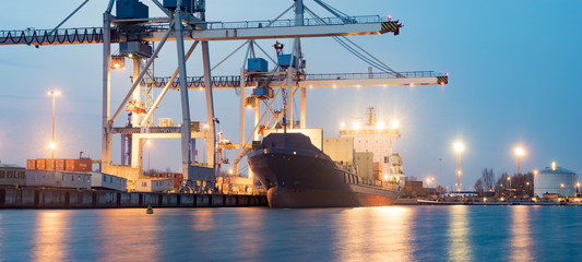 A large cargo ship docked at a port with cranes loading containers at dusk, lights reflecting on the calm water.
