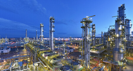 A large industrial oil refinery is brightly lit at dusk, with numerous towers, tanks, and pipes stretching across the complex under a blue sky.