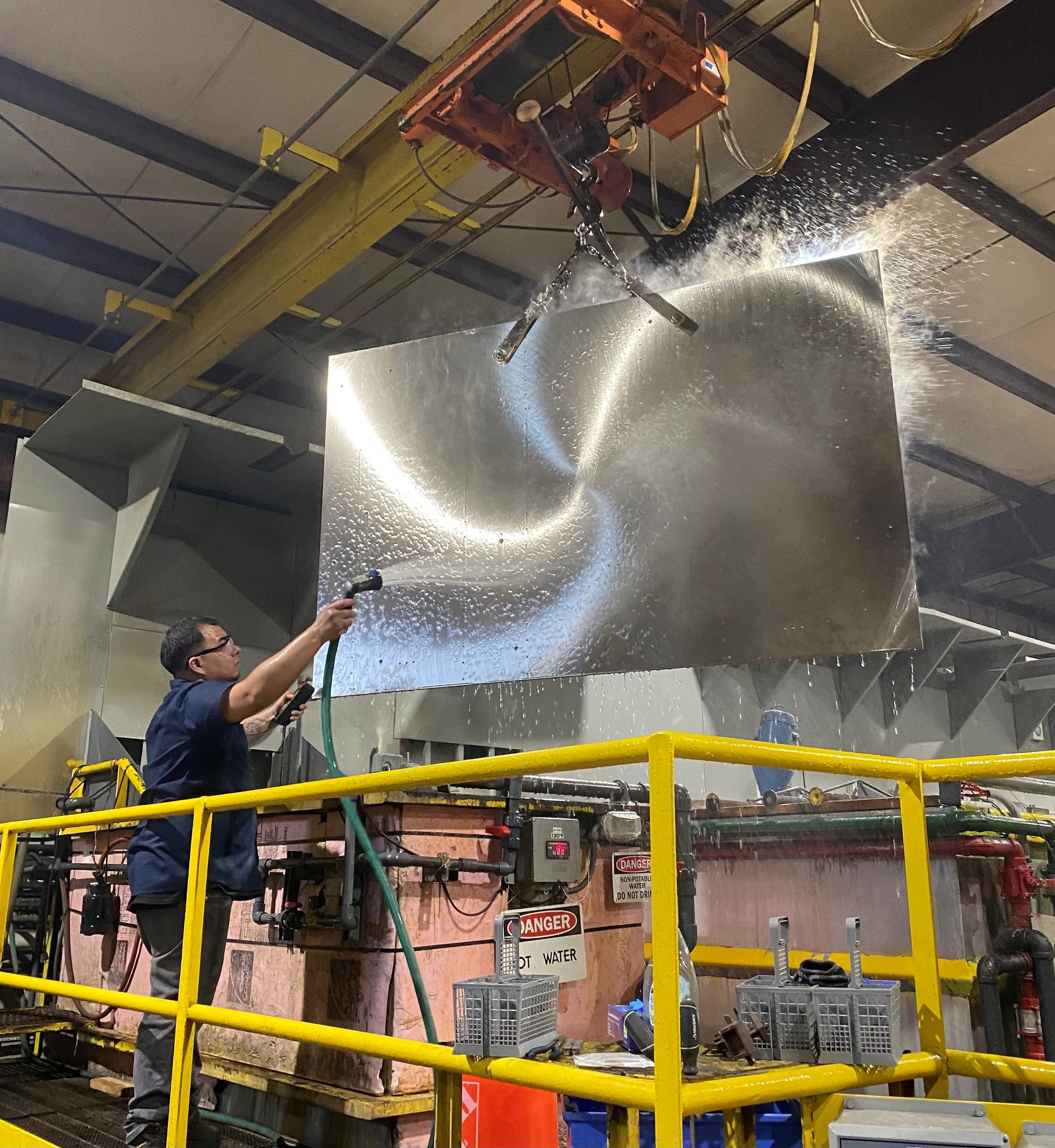 A worker sprays water on a large sheet of metal suspended by a crane in an industrial facility, with machinery and yellow railings surrounding the area.