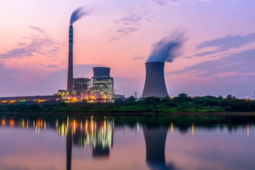 A power plant with smokestacks and cooling towers releases smoke into the evening sky, reflected in a calm body of water with greenery in the foreground.