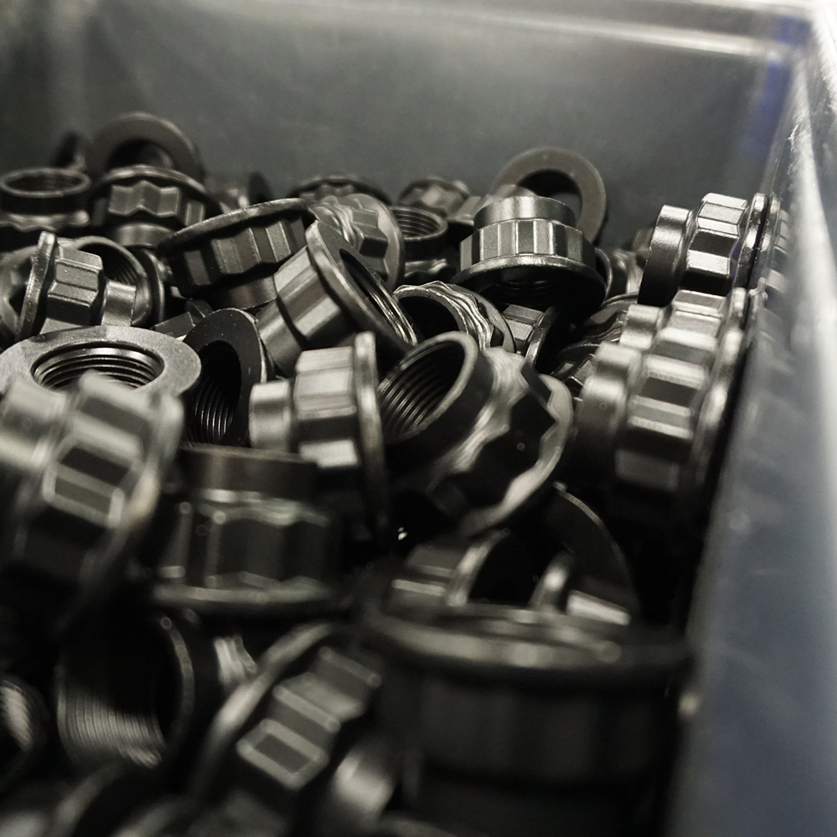 A close-up view of a bin filled with many black plastic threaded caps or nuts, arranged in a somewhat disorganized pile.
