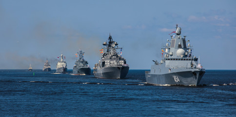 A fleet of navy warships sails in formation on open water under a clear blue sky, with several vessels visible in the distance.