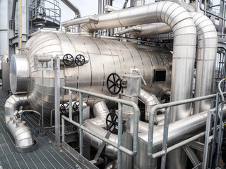 Large industrial metal tank with multiple pipes and control valves, surrounded by metal railings and platforms in a factory or processing plant setting.