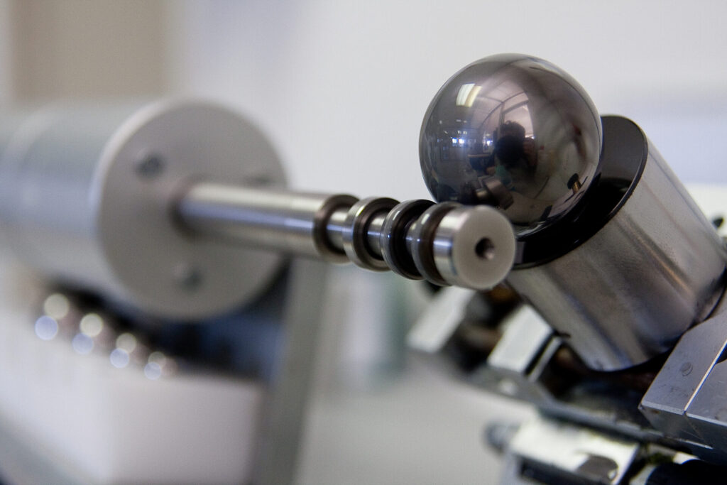 Close-up of a precision metal instrument with a shiny steel sphere mounted on a rod with coils, likely used in a scientific or industrial setting. The background is blurred, focusing attention on the device.