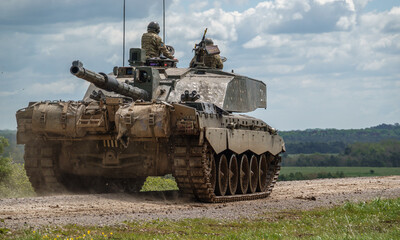 A military tank with soldiers on top drives on a dirt road in a rural area, with green fields and cloudy sky in the background.
