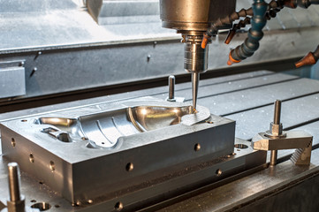 A CNC milling machine drills into a metal mold block, creating a detailed cavity design. Coolant hoses are visible, and metal shavings are scattered on the surface.