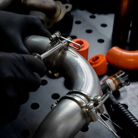 A person wearing black gloves uses a tool to adjust a clamp on a curved metal pipe with holes, with orange components nearby on a workbench.