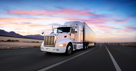 A white semi-truck drives on an empty highway at sunset, with mountains and a colorful sky in the background.