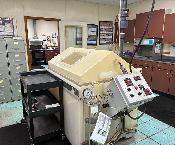 Large beige industrial machine with control panel and gauges in a lab, surrounded by file cabinets, a black cart, and brown cabinets. Lab equipment and office materials are visible in the background.