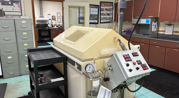 Laboratory room with a large beige machine featuring control panels and gauges, surrounded by cabinets, filing cabinets, charts, and a rolling cart on a blue tile floor.