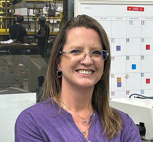 A woman with glasses and a purple shirt smiles in front of a whiteboard calendar. Industrial equipment and people working are visible in the background.