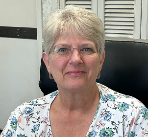 Smiling older woman with short gray hair and glasses sits in an office chair, wearing a white floral shirt.