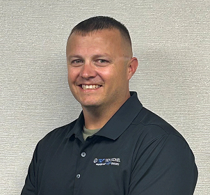 Smiling man with short hair wearing a dark polo shirt stands in front of a textured light-colored wall with his arms crossed.