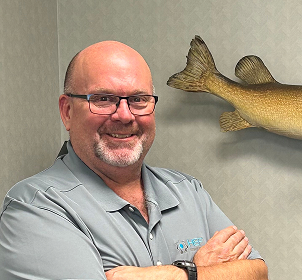 A man with glasses and a beard, wearing a light gray polo shirt, stands smiling with folded arms. A mounted fish tail is visible on the wall behind him.