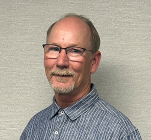 A man with glasses, a short beard, and a mustache is smiling slightly. He is wearing a blue and white striped collared shirt and is standing in front of a light textured wall.