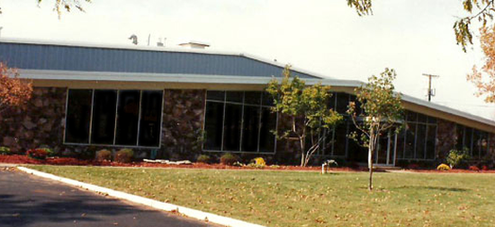 A single-story building with large windows and stone walls sits behind a green lawn with small trees and scattered autumn leaves under a clear sky.