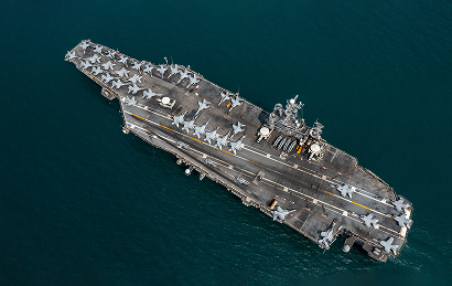 Aerial view of an aircraft carrier at sea with numerous fighter jets and helicopters positioned on the flight deck.