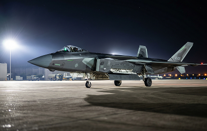 A modern stealth fighter jet with angular design is parked on an airfield at night under bright lights, with a row of buildings and runway lights visible in the background.