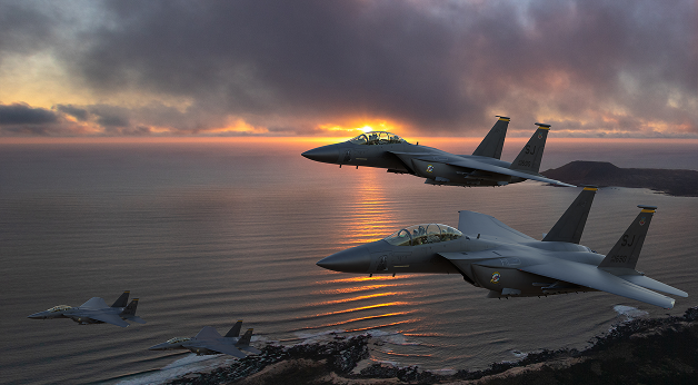 Four fighter jets fly in formation over a coastline at sunset, with dramatic clouds and the sun partly visible near the horizon, reflecting on the ocean below.