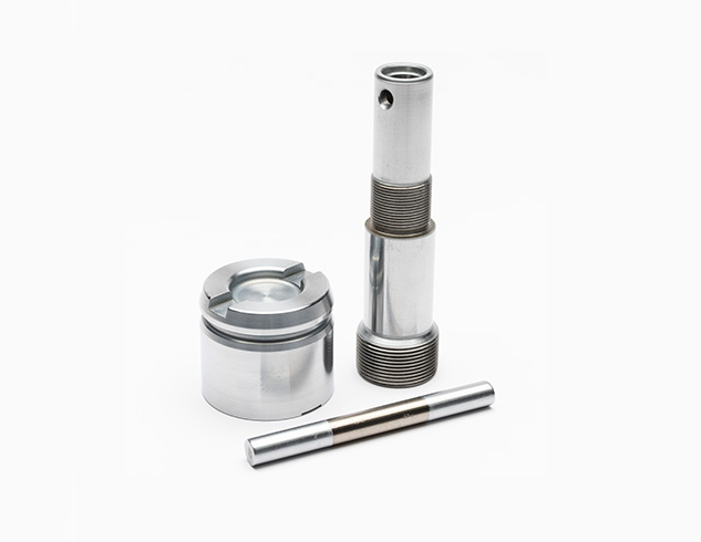 Three machined metal parts on a white background: a cylindrical piece with threads, a round piston-like component, and a slim, smooth rod lying in front.