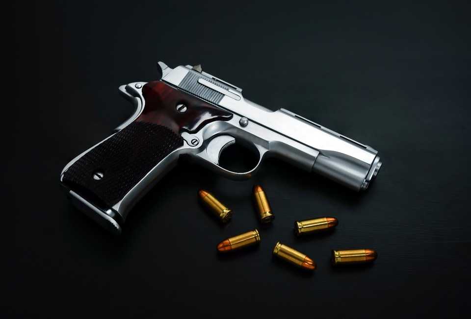 A silver handgun with a dark grip lies on a black surface next to six gold-colored bullets, arranged loosely around the firearm.