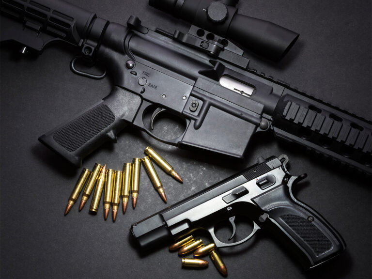 A black rifle and a black handgun are displayed on a dark surface, with scattered bullets placed nearby.