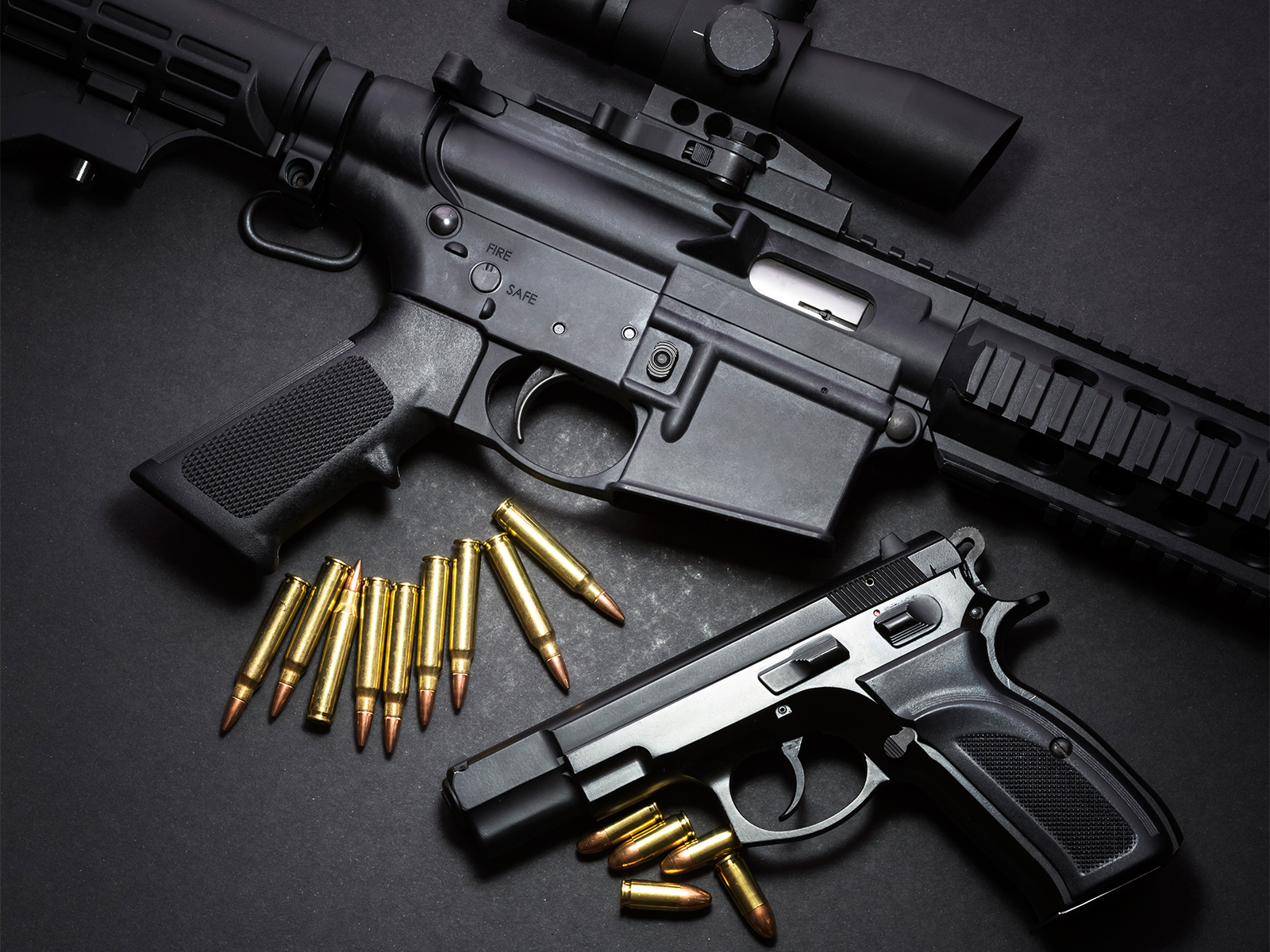 A black rifle and a black handgun are displayed on a dark surface, with scattered bullets placed nearby.