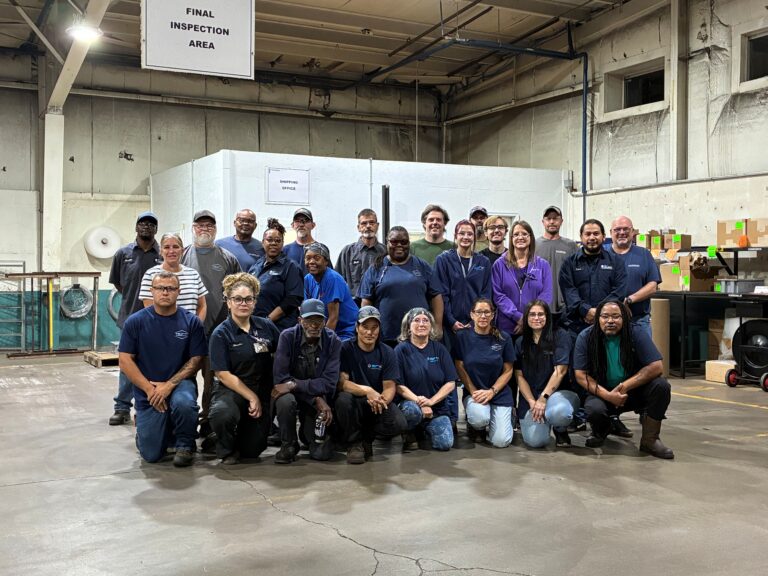 A group of about twenty-five people pose and smile inside an industrial warehouse under a sign labeled Final Inspection Area. They wear casual or company uniforms and stand and kneel in two rows.