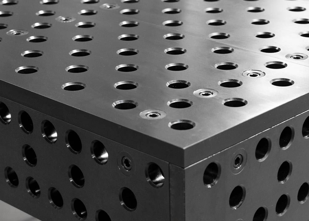 Close-up of two black metal plates with evenly spaced round holes, stacked at a right angle. The holes line up in a grid pattern, creating a geometric, industrial look.