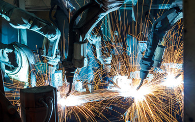 Robotic arms weld metal in a factory, creating bright sparks that illuminate the dark industrial environment. Automated machinery is used for precision manufacturing and assembly.
